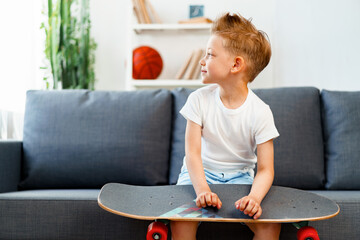 Little boy sitting on sofa at home and holding skateboard