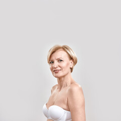 Fototapeta premium Portrait of cheerful beautiful middle aged woman in white bra looking at camera, posing isolated against grey background