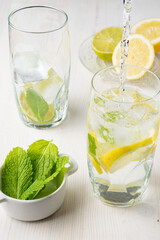 Top view of water falling into glass glass with half lemons on plate and mint leaves, on white table, in vertical