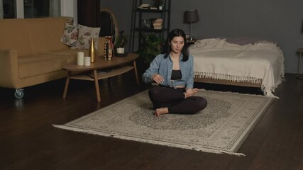 Relaxed young Asian woman in sportswear is meditating in lotus position sitting on floor with closed eyes at home enjoying meditation with hands in namaste.