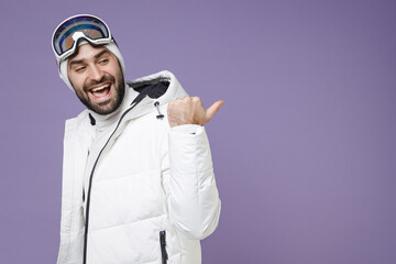 Side view of funny skier man in white windbreaker jacket ski goggles mask pointing thumb aside spend extreme weekend winter in mountains isolated on purple background. People lifestyle hobby concept.