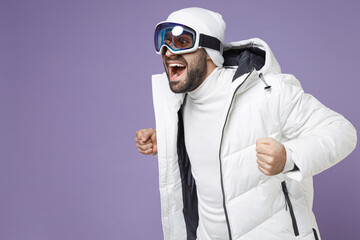 Surprised skier man in white windbreaker jacket ski goggles mask clenching fists looking aside spend extreme weekend winter in mountains isolated on purple background. People lifestyle hobby concept.