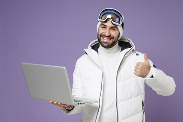 Funny skier man in warm white windbreaker jacket ski goggles mask work on laptop pc computer showing thumb up spend weekend in mountains isolated on purple background. People lifestyle hobby concept. © ViDi Studio