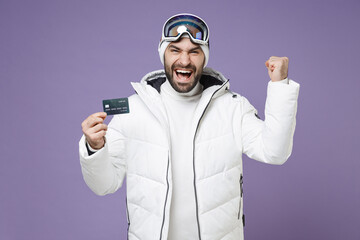Happy skier man in warm white windbreaker jacket ski goggles mask hold credit bank card doing...