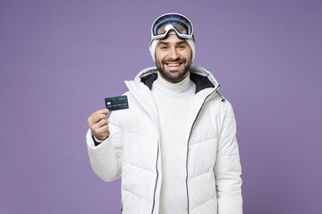 Funny skier man in warm white windbreaker jacket ski goggles mask hold credit bank card spend extreme weekend winter season in mountains isolated on purple background. People lifestyle hobby concept.