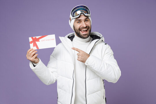 Cheerful skier man in white windbreaker jacket ski goggles mask point index finger on gift certificate spend weekend winter in mountains isolated on purple background. People lifestyle hobby concept.