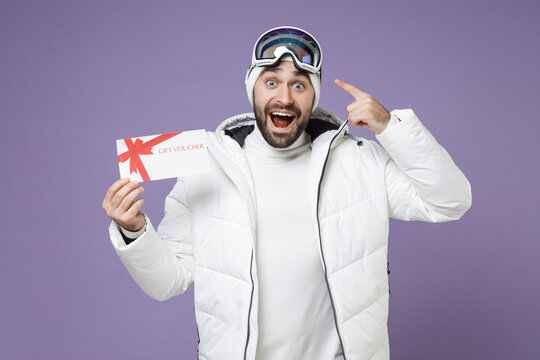 Shocked skier man in white windbreaker jacket pointing finger on ski goggles mask hold gift certificate spend weekend winter in mountains isolated on purple background. People lifestyle hobby concept.