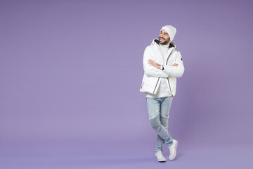 Full length of smiling man in warm white padded windbreaker jacket hat holding hands crossed looking aside isolated on purple background studio portrait. People lifestyle cold winter season concept.