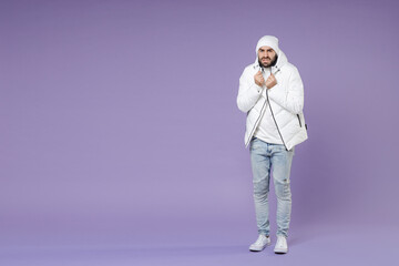 Full length of worried displeased freezing man in white padded windbreaker jacket hat looking camera isolated on purple color background studio portrait. People lifestyle cold winter season concept. © ViDi Studio