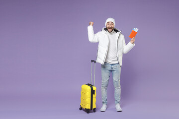 Full length of joyful traveler tourist man in white jacket hat hold passport ticket suitcase doing winner gesture isolated on purple background. Passenger traveling on weekends. Air flight concept.