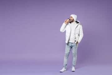 Full length funny man in warm white windbreaker jacket hat hold paper cup coffee tea drinking looking aside isolated on purple background studio portrait. People lifestyle cold winter season concept.