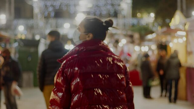 A view from the back on a brunette woman in a big red fur coat and a surgical covid-19 face mask wandering around European Christmas market. Lights, decorations on background. High-quality 4k video.