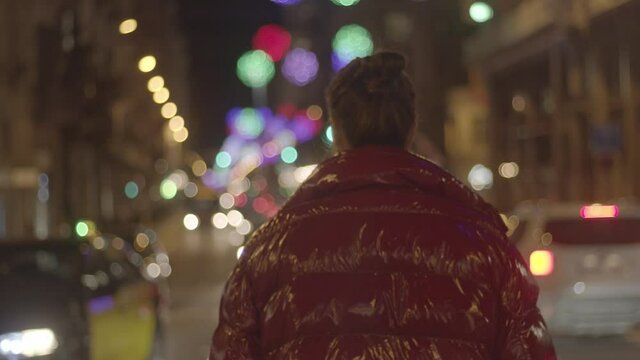 Caucasian girl on a winter urban night. In a red jacket, white sweater and a medical face mask. Blurry Christmas lights cars and taxis in the background. Medium close-up, 4K high-quality video footage
