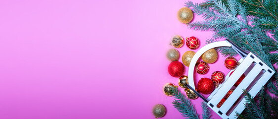 New Year's Christmas tree decorations of red and gold color lie in a chaotic manner on a pink background among fir branches covered with snow, and a wooden box stands nearby. 