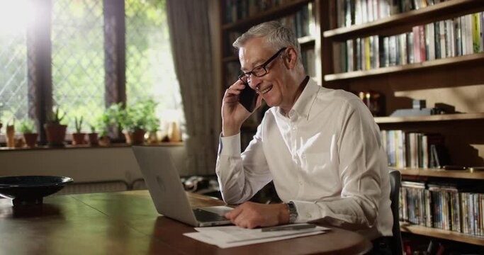 4K Smiling casual man making friendly phone call while working on laptop at home.