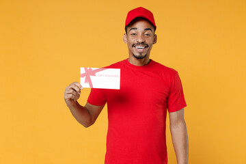 Delivery employee african man 20s in red cap blank print t-shirt uniform workwear work courier dealer service concept hold mockup gift certificate voucher coupon isolated on yellow background studio.