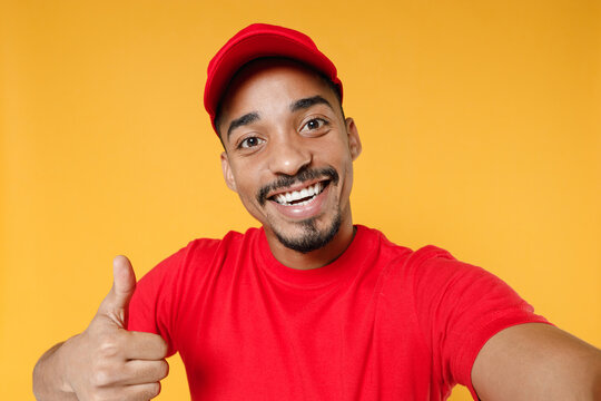 Close up fun delivery employee african man in red cap blank print tshirt uniform workwear work courier dealer service concept doing selfie shot on mobile phone isolated on yellow background studio