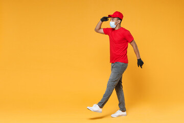 Full length delivery employee african man in red cap t-shirt face mask gloves uniform work courier dealer service on quarantine coronavirus covid-19 virus concept isolated on yellow background studio.