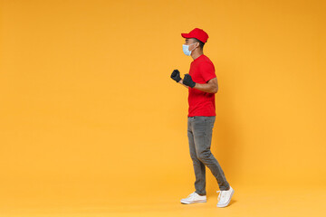 Full length delivery employee african man in red cap t-shirt face mask gloves uniform work courier dealer service on quarantine coronavirus covid-19 virus concept isolated on yellow background studio.