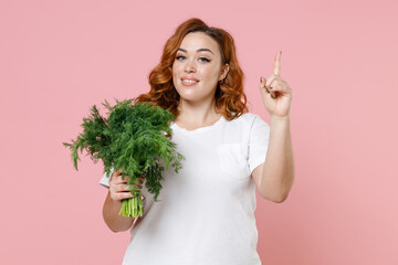 Smiling young redhead plus size body positive woman 20s in white t-shirt hold bunch of fresh greenery dill pointing index finger up looking camera isolated on pastel pink background studio portrait.