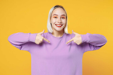 Smiling fun young blonde caucasian woman 20s wearing casual basic purple shirt pointing index finger on sweatshirt looking camera toothy smile isolated on yellow color background studio portrait. - Powered by Adobe