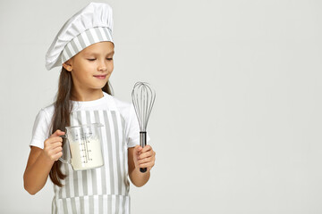 little girl in chef hat and apron with flour in measuring cup and whisk. child dreams of becoming a chef. copy space