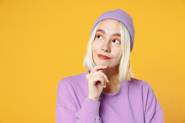 Pensive dreamful young blonde caucasian woman 20s bob haircut bright makeup wearing basic purple shirt violet beanie hat prop up chin look aside isolated on yellow color background studio portrait.