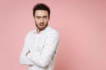 Fototapeta premium Shocked worried scared young bearded man 20s wearing basic casual white shirt standing holding hands crossed keeping mouth open looking camera isolated on pastel pink color background studio portrait.