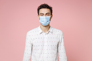 Young bearded man 20s wearing basic casual white shirt sterile face mask to safe from coronavirus virus covid-19 during pandemic quarantine isolated on pastel pink color background studio portrait.