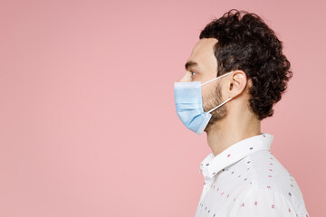 Side view of young bearded man 20s in basic white shirt sterile face mask to safe from coronavirus virus covid-19 during pandemic quarantine isolated on pastel pink color background studio portrait.