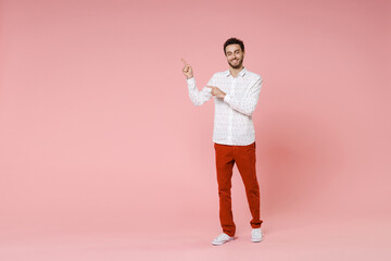 Full length of smiling young bearded man 20s wearing basic casual white shirt standing pointing index fingers aside on mock up copy space isolated on pastel pink color wall background studio portrait.