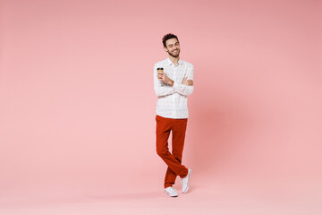 Fototapeta na wymiar Full length of smiling young bearded man 20s wearing basic casual white shirt standing hold paper cup of coffee or tea hold hands crossed isolated on pastel pink color wall background studio portrait.