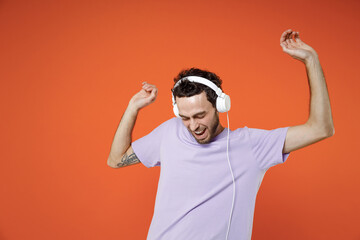 Naklejka premium Cheerful laughing funny young bearded man 20s wearing casual basic violet t-shirt listening music with headphones dancing rising hands isolated on bright orange color wall background studio portrait.