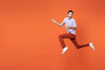 Fototapeta na wymiar Full length side view of shocked screaming young bearded man 20s wearing casual violet t-shirt jumping pointing index fingers aside isolated on bright orange color wall background studio portrait.