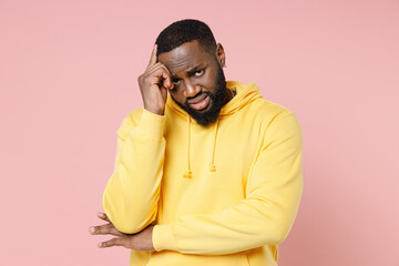 Confused worried young african american man 20s wearing casual basic yellow streetwear hoodie standing put hand on head looking camera isolated on pastel pink color wall background studio portrait.