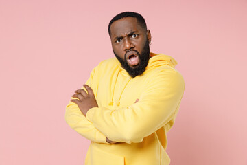 Side view of shocked worried young african american man 20s wearing casual basic yellow streetwear hoodie holding hands crossed looking camera isolated on pastel pink color background studio portrait.