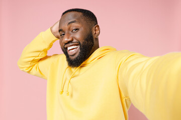 Close up of smiling young african american man 20s wearing casual basic yellow streetwear hoodie doing selfie shot on mobile phone put hand on head isolated on pastel pink background studio portrait.