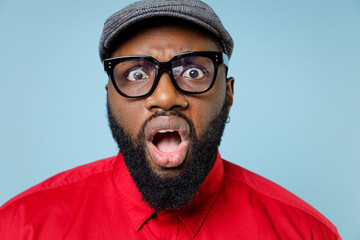 Close up of shocked worried young bearded african american man 20s wearing casual red shirt cap eyeglasses keeping mouth open looking camera isolated on pastel blue color background studio portrait.
