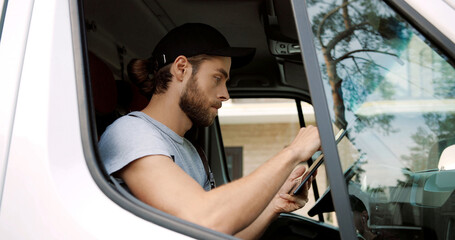 Side view of Caucasian handsome male courier sitting on front seat in van tapping and typing on tablet device delivering orders. Close up of delivery man in uniform using gadget. Work concept