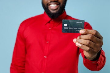 Cropped image of smiling funny young bearded african american man 20s wearing casual red shirt standing hold in handcredit bank card isolated on pastel blue color wall background studio portrait.