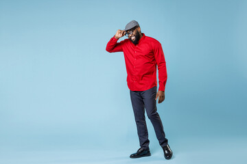 Full length of smiling young bearded african american man 20s wearing casual red shirt hold cap eyeglasses standing and looking camera isolated on pastel blue color wall background studio portrait.