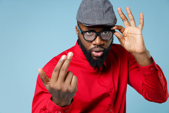 Young bearded african american man 20s wearing casual red shirt cap eyeglasses standing calling with fingers gesture like says come to me isolated on pastel blue color wall background studio portrait.