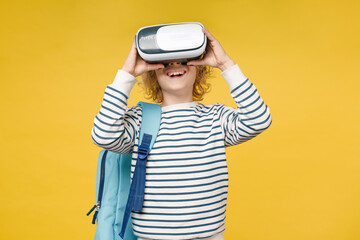 Cheerful little male kid teen boy 10s years old wearing striped sweatshirt glasses backpack watching in vr headset gadget isolated on yellow color background, child studio portrait. Education concept.