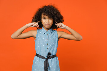 Worried little african american kid girl 12-13 years old in casual denim dress covering ears with fingers isolated on orange color background children studio portrait. Childhood lifestyle concept.