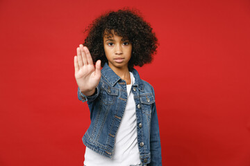 Serious little african american kid girl 12-13 years old in casual denim jacket showing stop gesture with palm isolated on red color background children studio portrait. Childhood lifestyle concept.