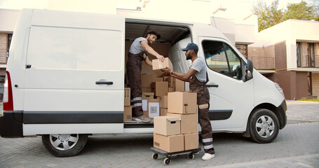 Young Caucasian delivery man giving boxes from van to African American colleague standing on street...