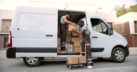 Young mixed-races delivery men coworkers taking boxes from van putting on cart standing on street outdoor delivering shipment. Male post office workers with shopping package. Courier concept © VAKSMANV