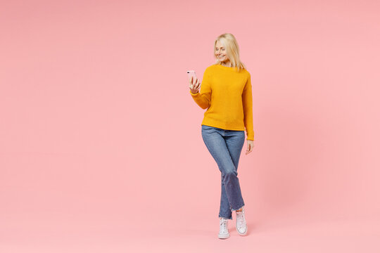 Full Length Of Smiling Elderly Gray-haired Blonde Woman Lady 40s 50s Years Old In Yellow Casual Sweater Using Mobile Cell Phone Typing Sms Message Isolated On Pink Color Background Studio Portrait.