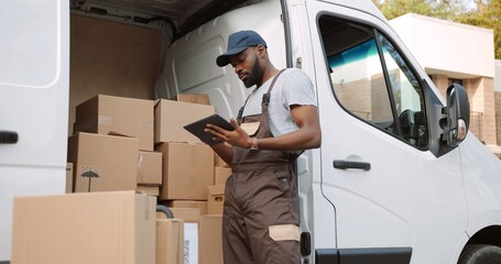 Young handsome African American delivery man typing and tapping on tablet device standing near van...