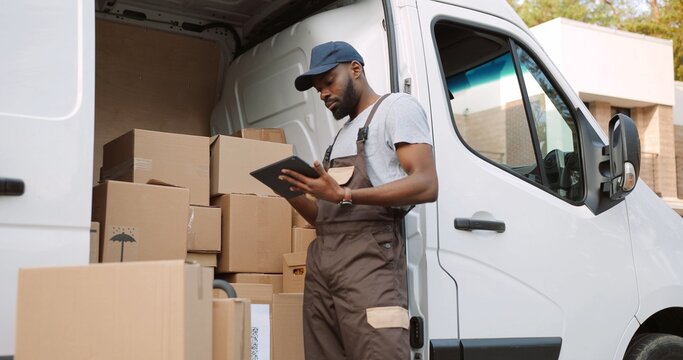 Young handsome African American delivery man typing and tapping on tablet device standing near van on street with many ordered boxes. Male postman courier with shopping package. Shipment concept